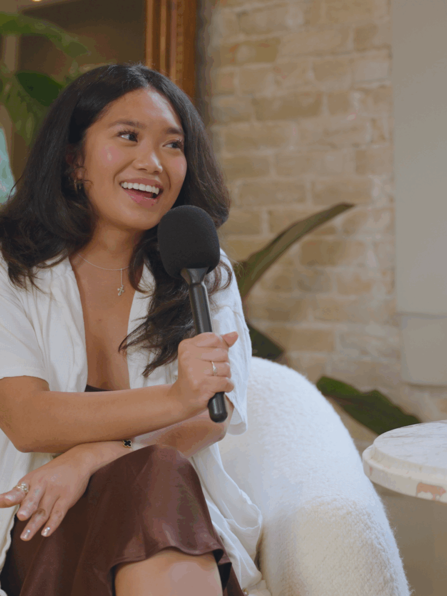 A woman with long dark hair smiles while sitting in a white chair, holding a microphone. She is wearing a white top and brown skirt. There is a round table with a plant and a cup beside her indoors with plants in the background.