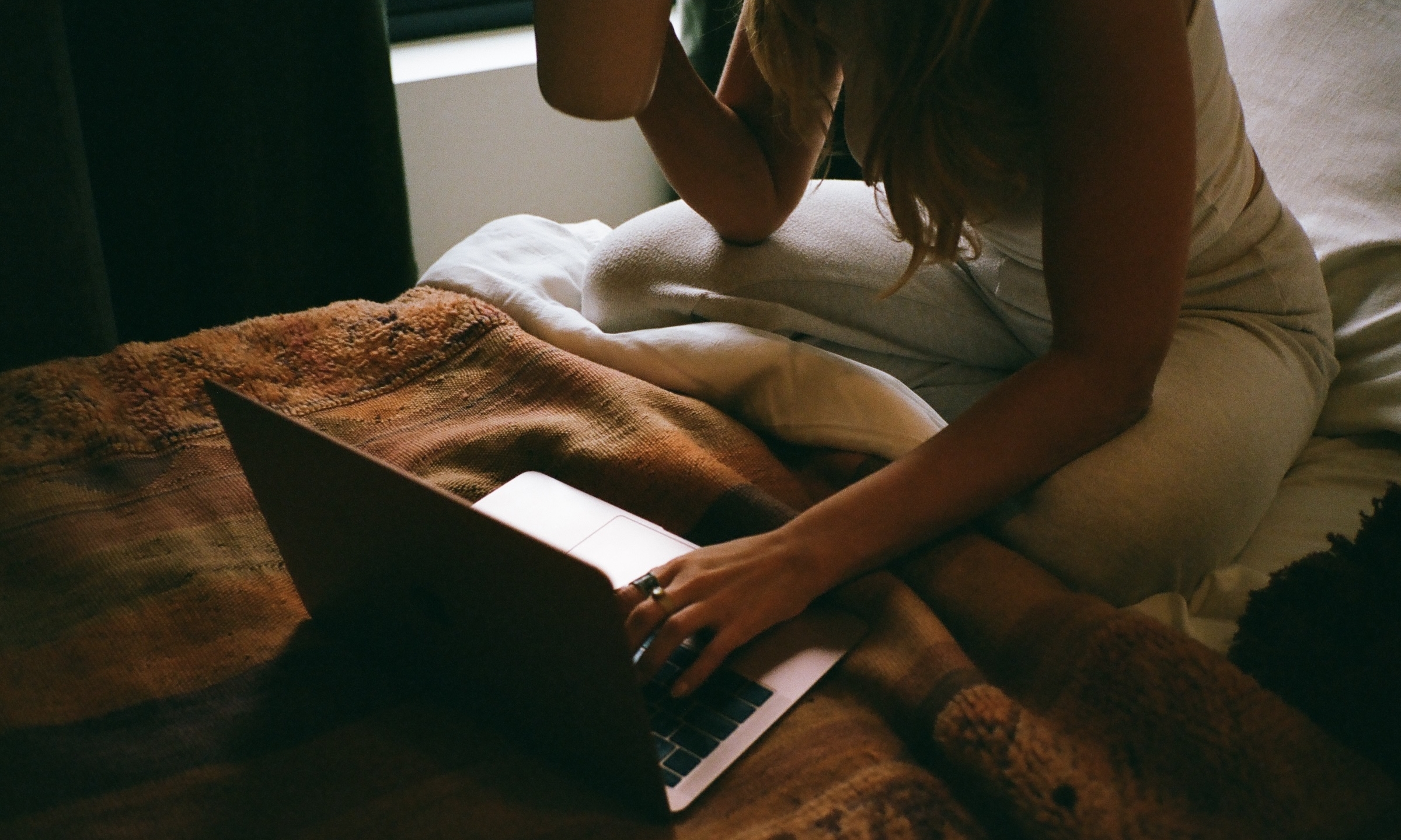 A person sits on a bed with a brown blanket, holding a mug in one hand and typing on a laptop with the other, next to a window letting in natural light.