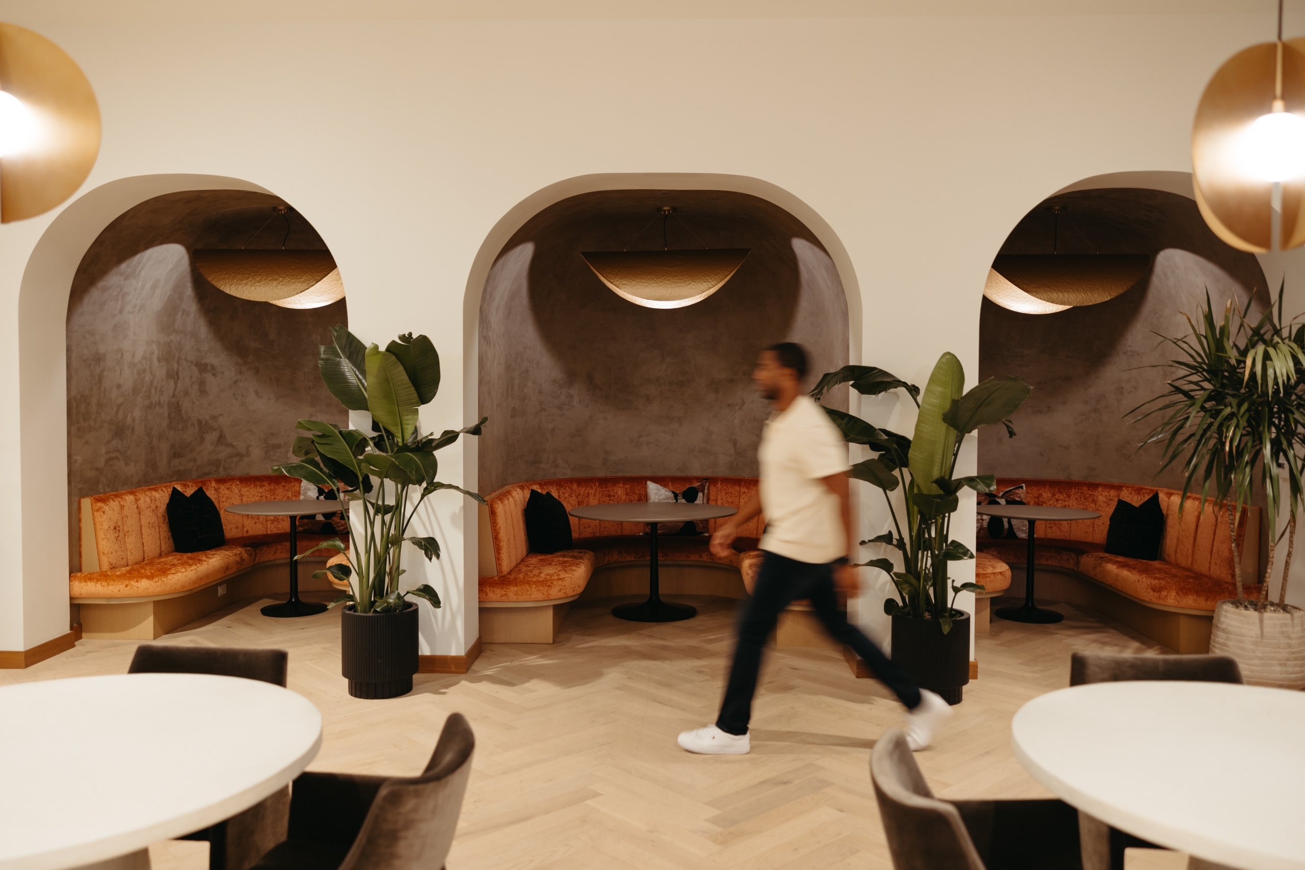 A blurred person walks past modern, cozy booth seating with orange cushions and potted plants in a stylish, well-lit lounge area with arched alcoves.