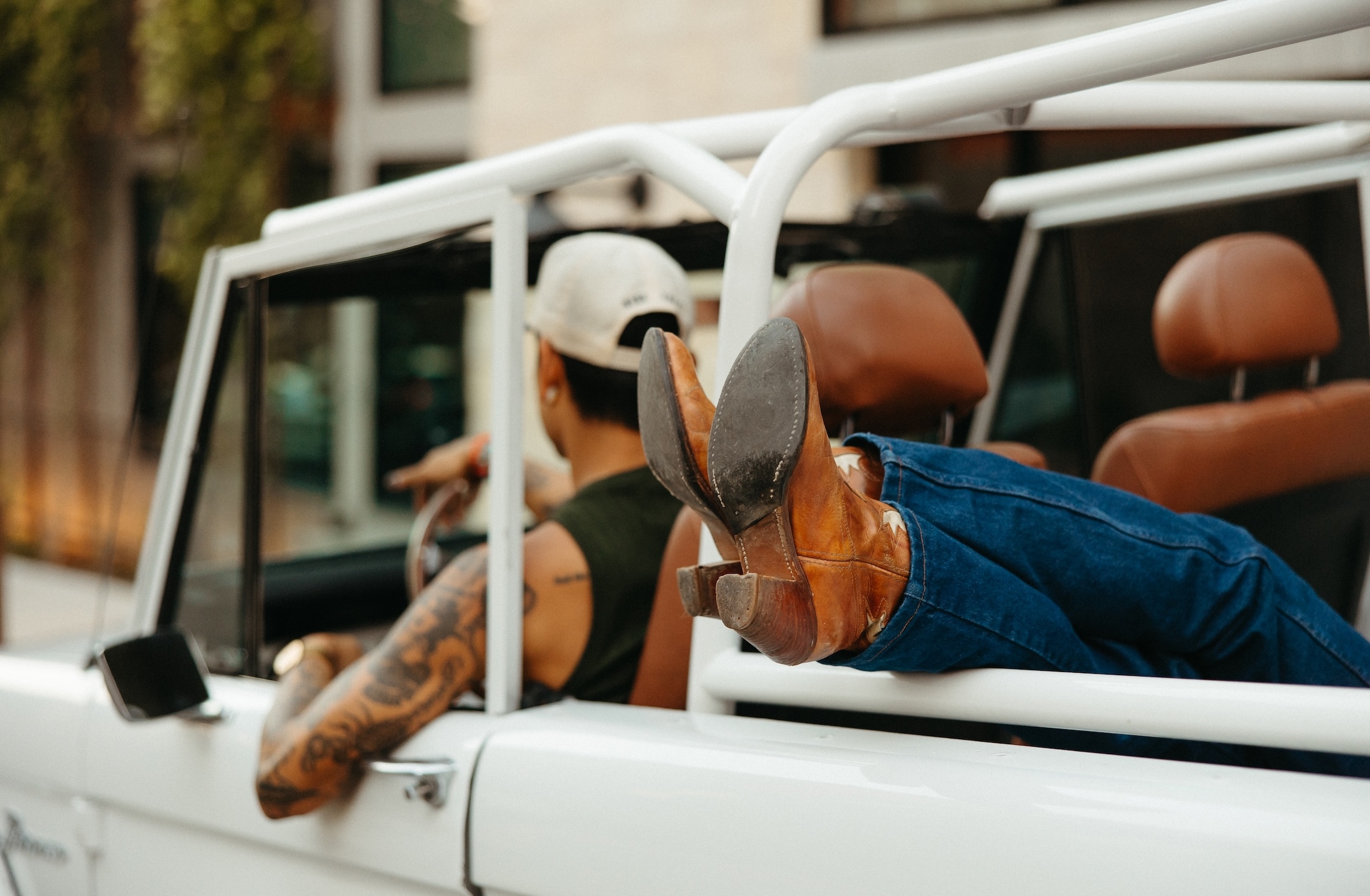 A person wearing jeans and cowboy boots relaxes with their feet out the side of a white LV Collective convertible. Seen from behind, they sport a white cap, show off arm tattoos, and hold the steering wheel.