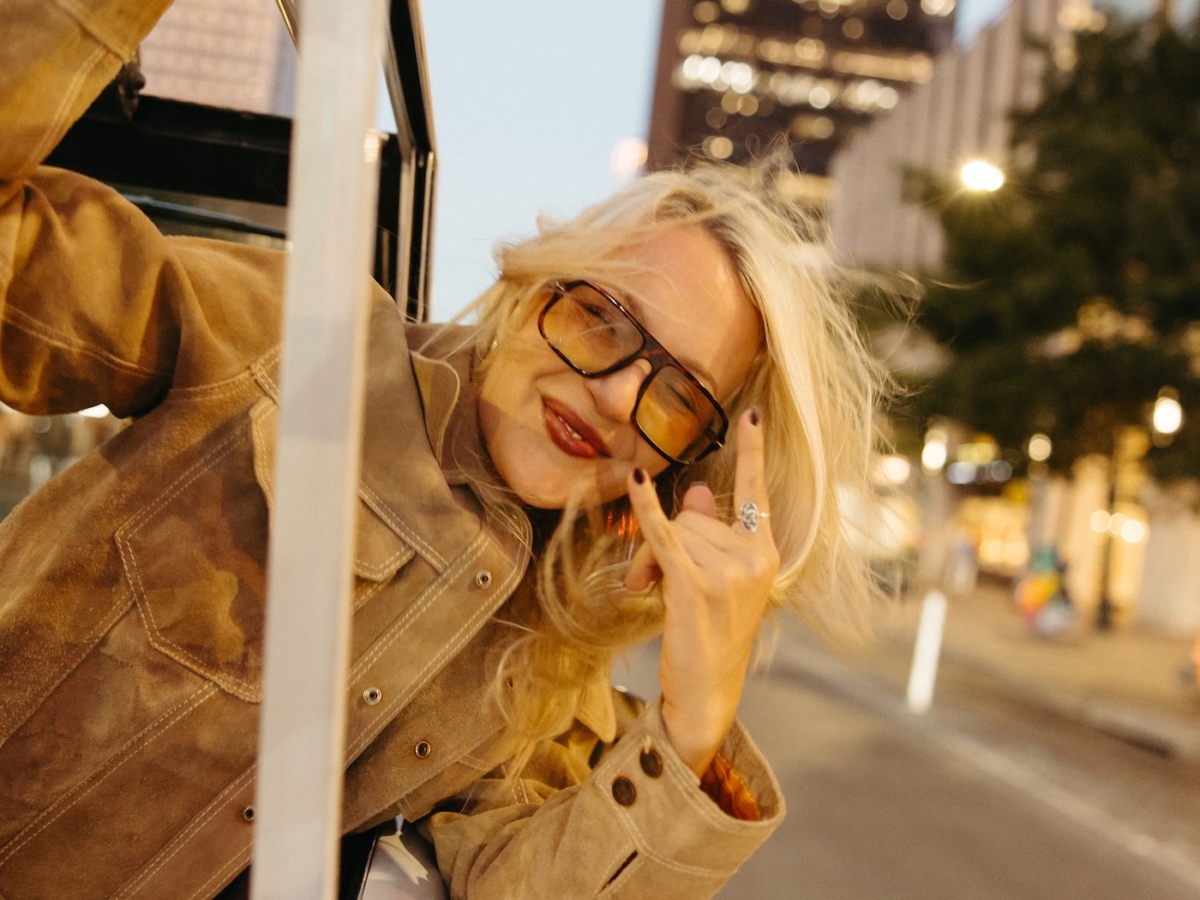 A woman with long blonde hair leans out of a car window, smiling and making a hand sign, wearing large sunglasses and a tan jacket, with city buildings and street lights reflecting the vibrant energy of LV Collective in the background.