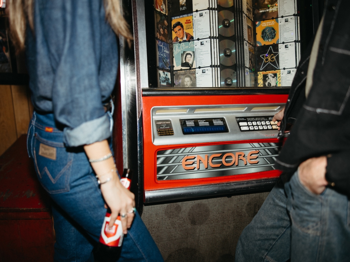 Two people stand near a vintage red Encore jukebox, one holding a drink bottle. The jukebox displays various music albums, and both individuals wear casual denim clothing—a classic LV Collective vibe.