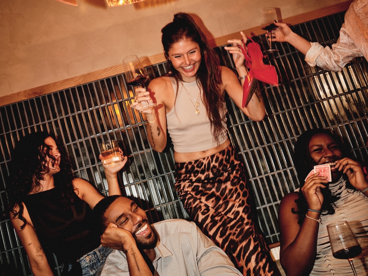 A group of friends enjoy a lively night out; one woman stands on a seat holding a drink and red heels, while others—part of the LV Collective—sit laughing, playing cards, and holding wine glasses around a table under warm lighting.