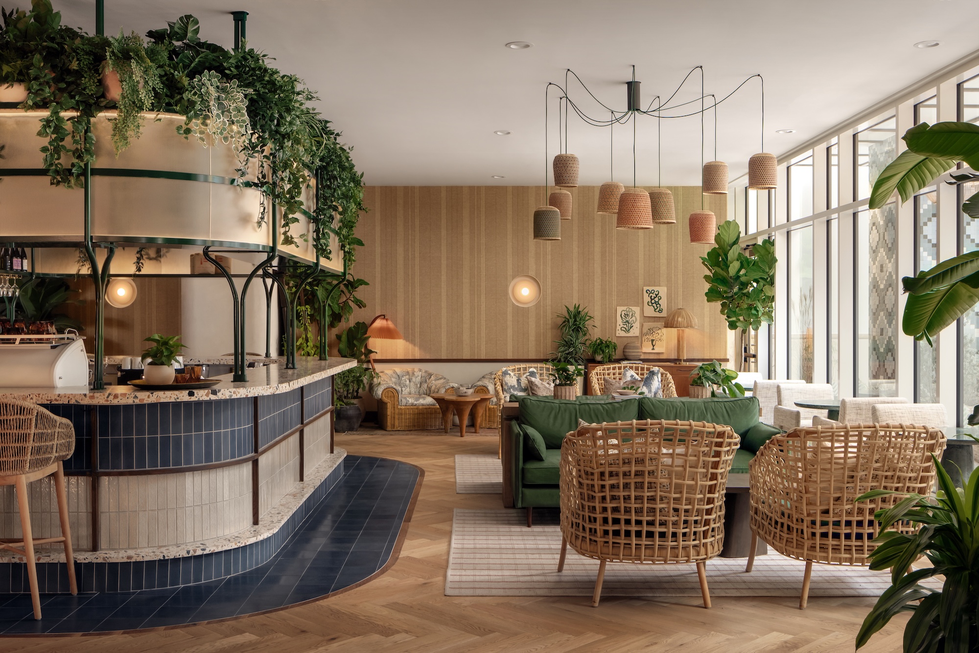 A stylish cafe with rattan chairs, green sofa, hanging plants, and warm lighting. The space features large windows, wooden floors, and cozy seating areas with natural decor and modern accents.