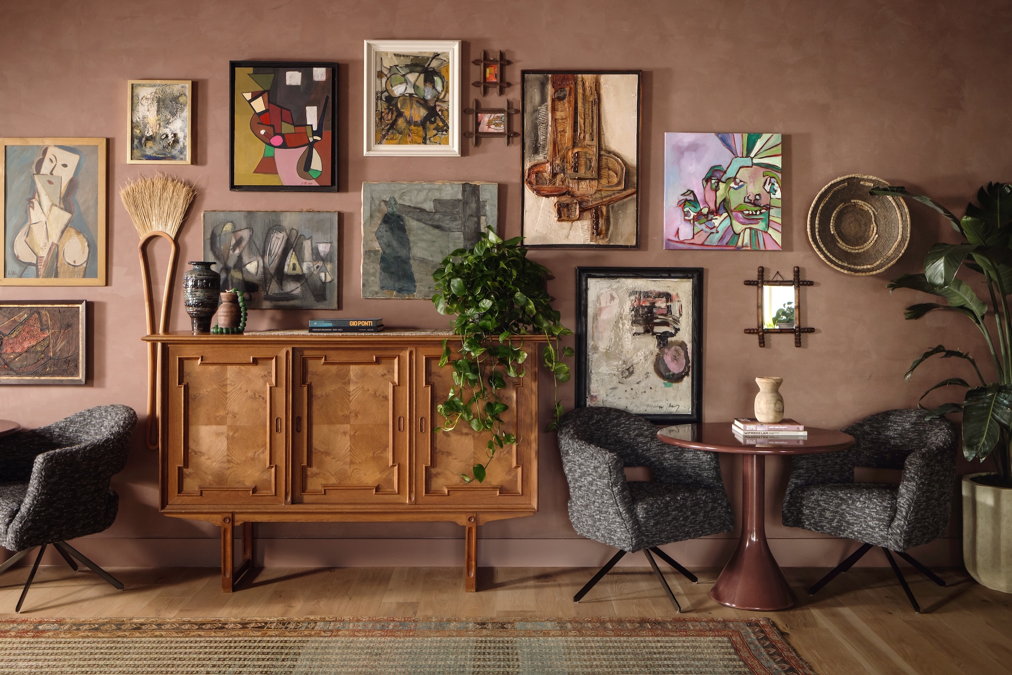 A cozy LV Collective room with a wooden sideboard, gray armchairs, a small round table, potted plants, and a mauve wall decorated with assorted paintings, masks, baskets, and art pieces. A rug covers part of the wooden floor.