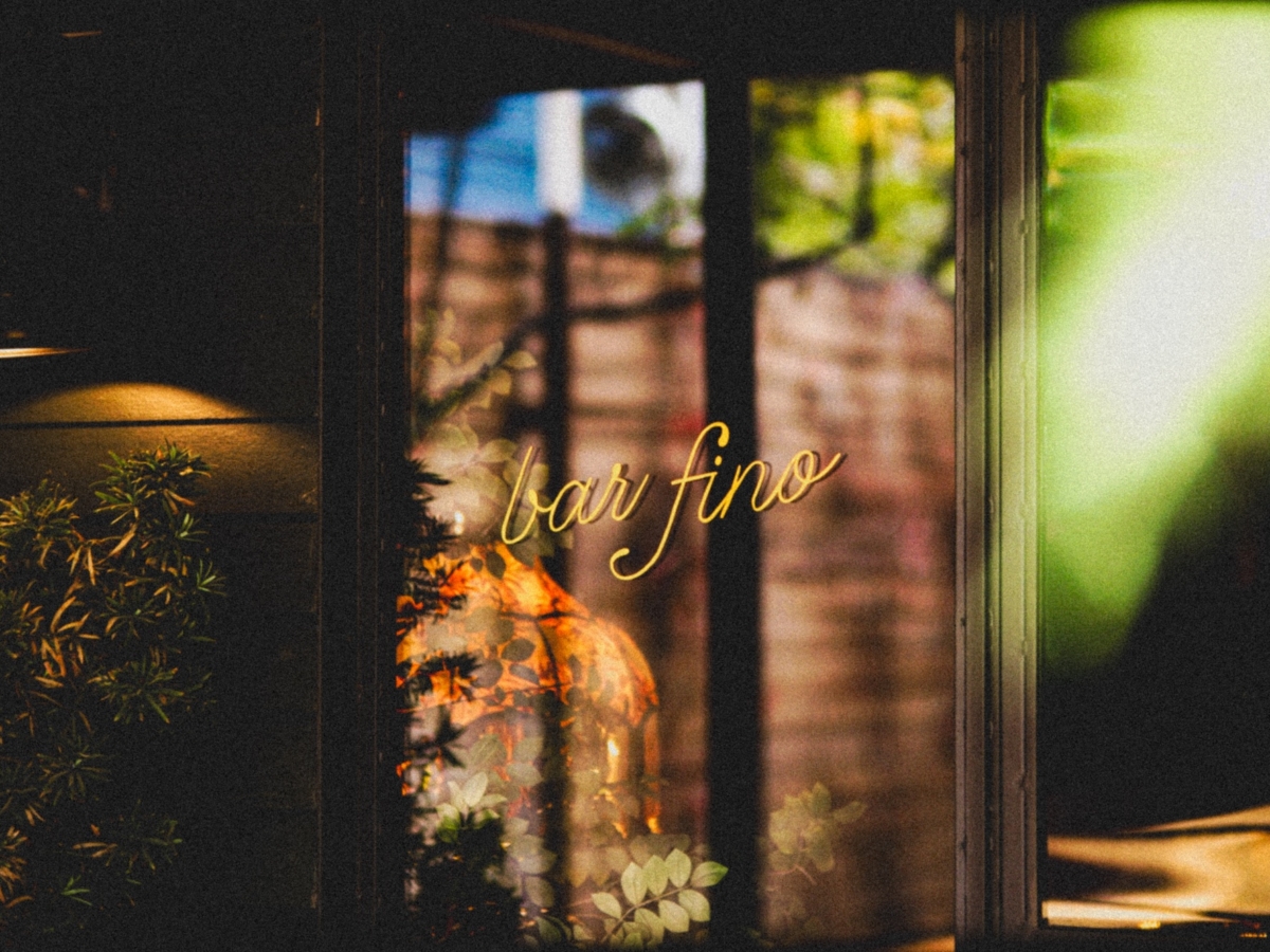 A softly lit window with the words bar fino written in elegant yellow script, reflecting plants and warm light from inside, creates a cozy and inviting atmosphere inspired by LV Collective’s signature style.