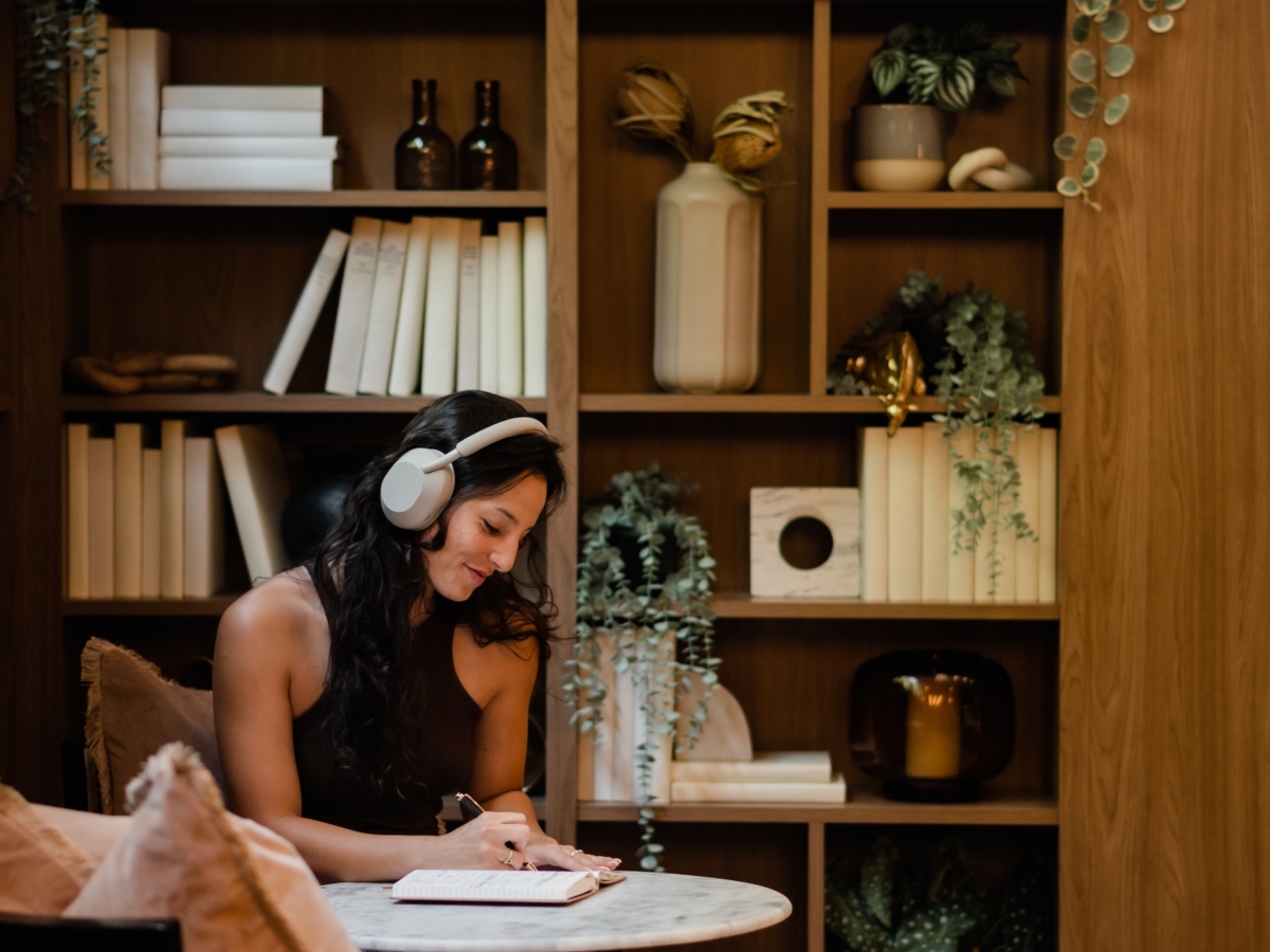 A woman wearing headphones sits at a round table in a cozy, plant-filled room by LV Collective, writing in a notebook. Behind her, a wooden bookshelf is decorated with books, greenery, and decorative objects.