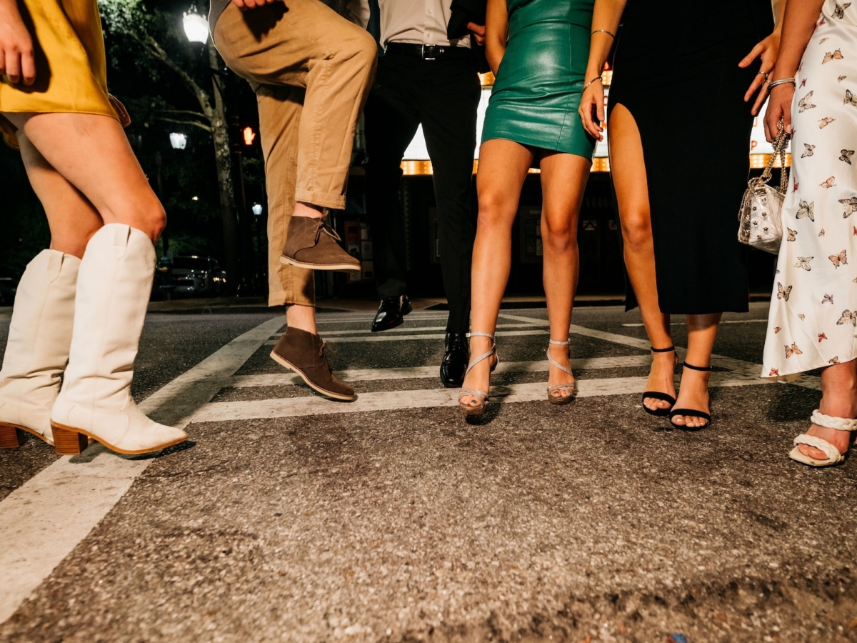Five people stand on a city street at night, their legs visible from the knees down. Each showcases stylish dresses, skirts, pants, and chic footwear—boots, heels, and loafers—reflecting the signature flair of the LV Collective.