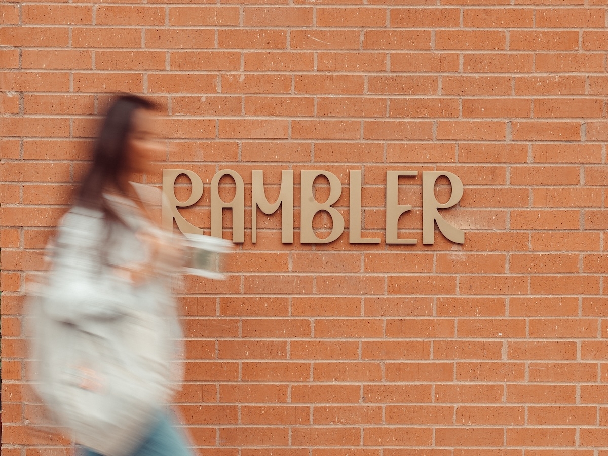 A person in motion walks past a red brick wall with the word RAMBLER displayed in large, modern letters—a signature touch by LV Collective. The person is blurred, carrying a drink and a bag.