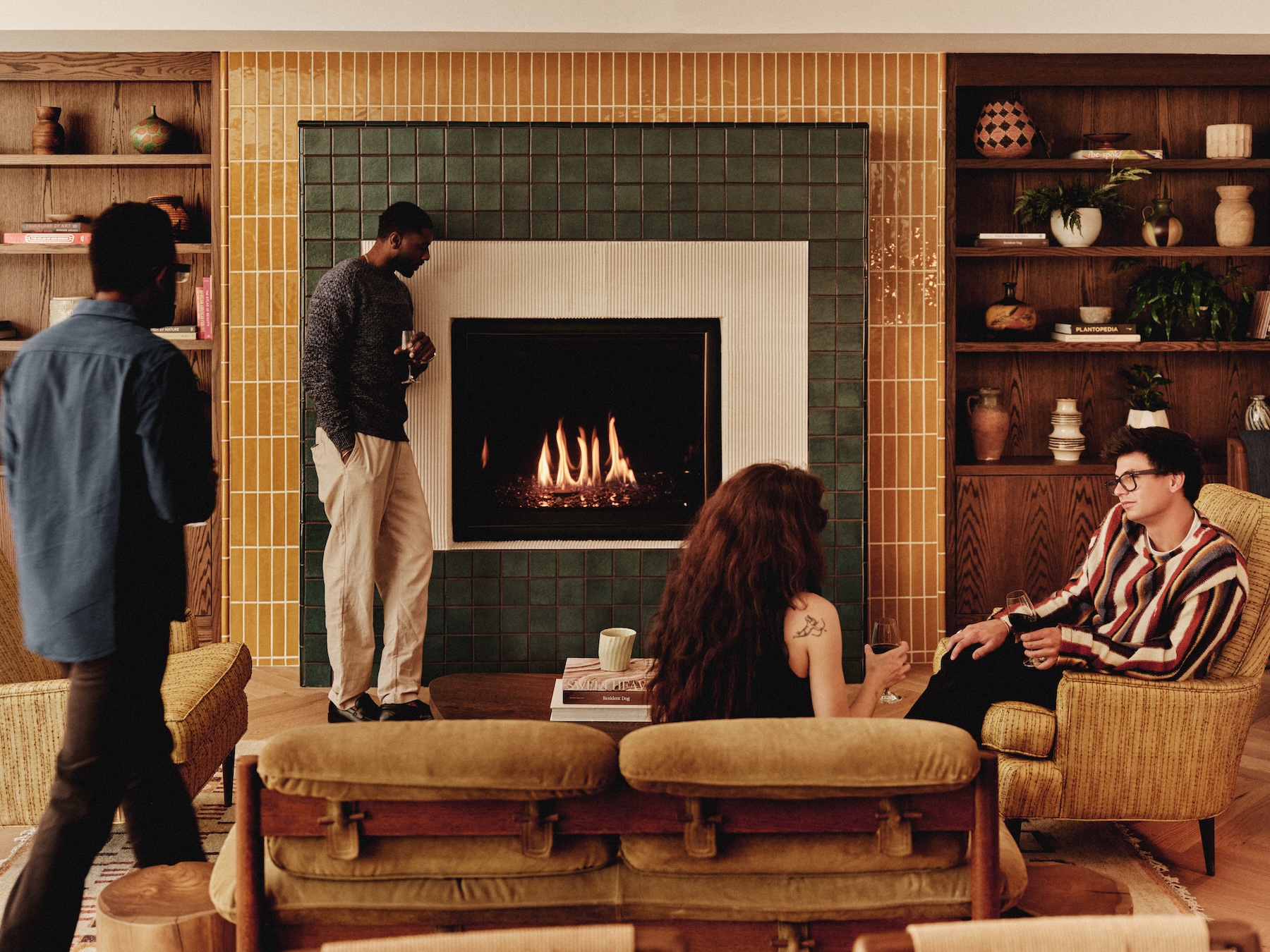 Four people gather in a cozy LV Collective living room with a lit fireplace, yellow armchairs, and wooden shelves adorned with plants and pottery. Two sit and chat, one stands by the fire, and another walks into the welcoming space.