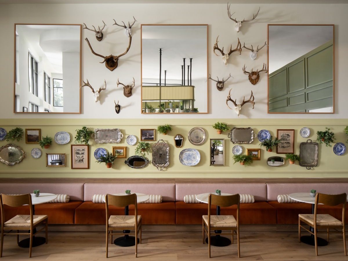 A stylish cafe interior with wooden chairs and cushioned bench seating. The wall displays mirrors, mounted antlers, decorative plates, trays, framed art, and small potted plants, creating an eclectic and cozy atmosphere.