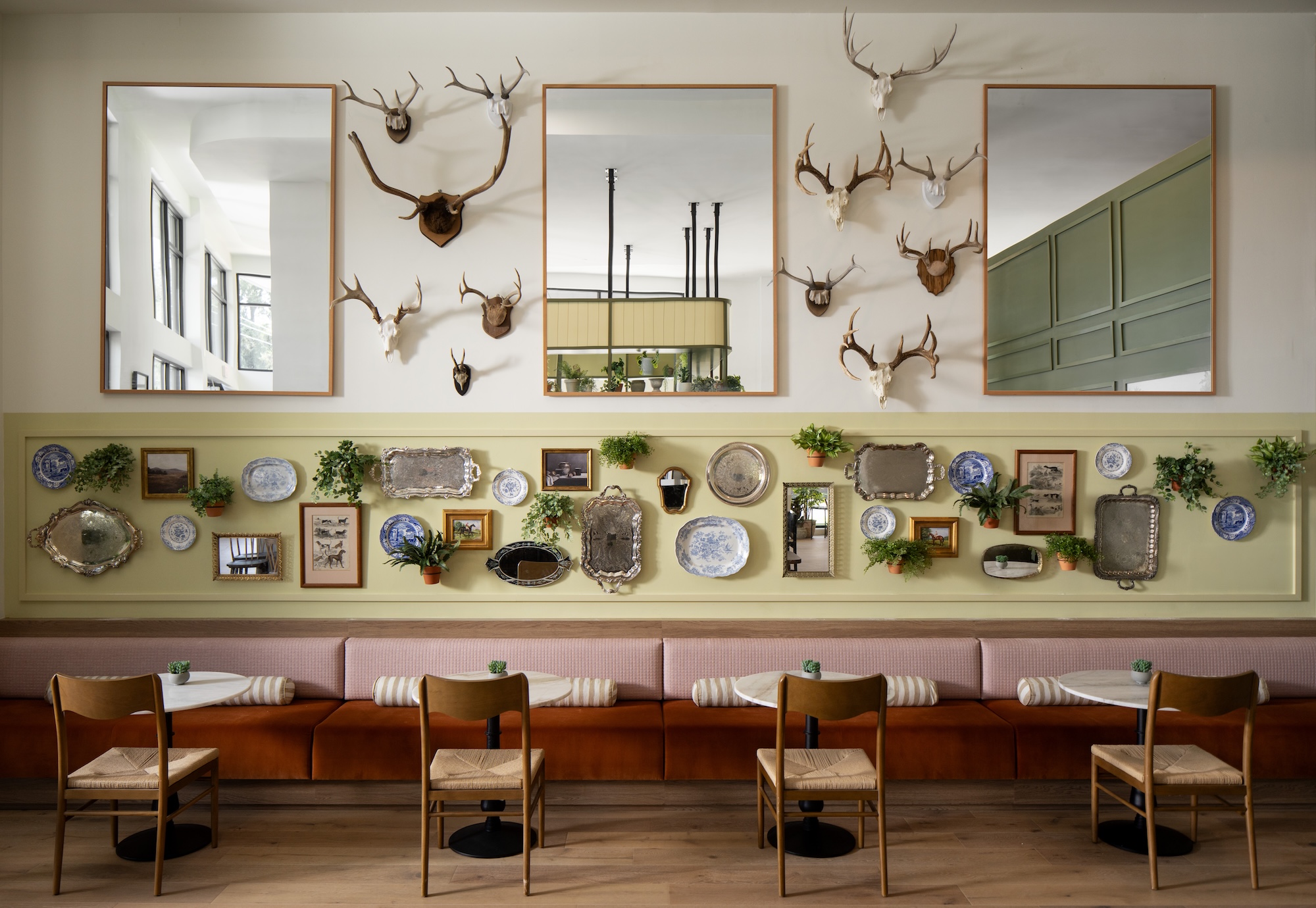 A stylish cafe interior with wooden chairs and cushioned bench seating. The wall displays mirrors, mounted antlers, decorative plates, trays, framed art, and small potted plants, creating an eclectic and cozy atmosphere.