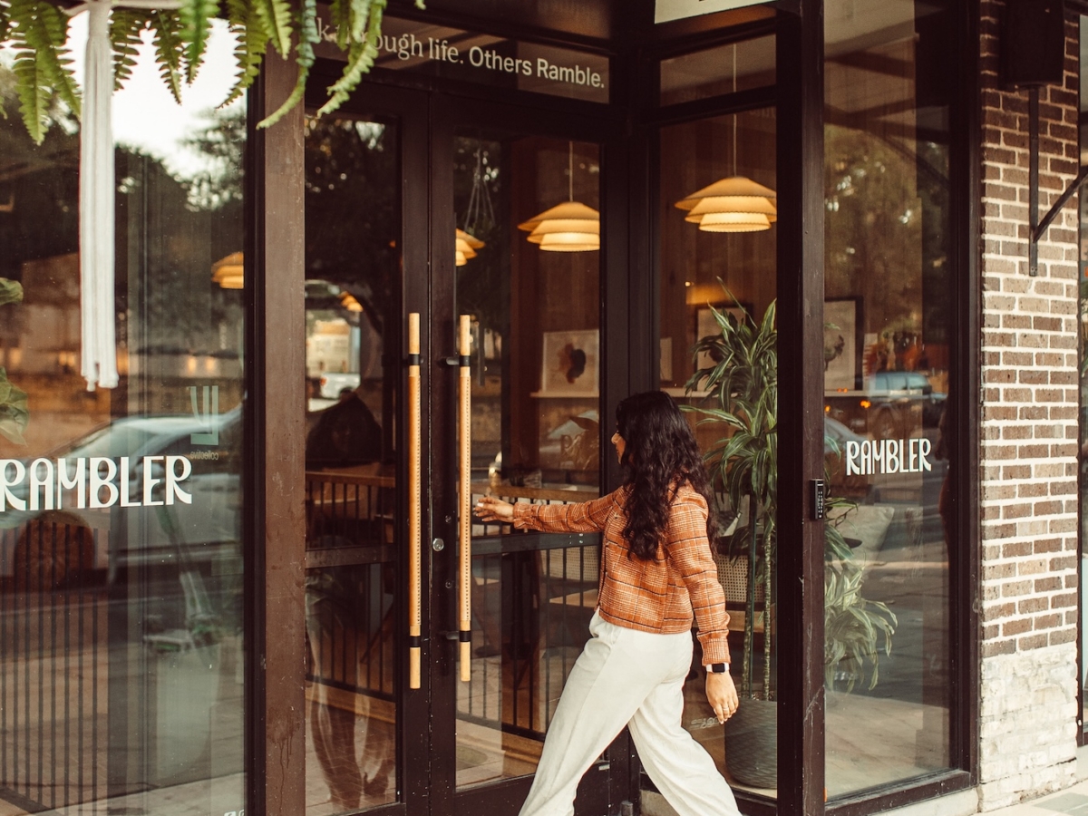 A person with long dark hair, wearing a brown jacket and light-colored pants, is opening the glass door to LV Collective’s café Rambler. Green plants hang overhead, and painted shapes brighten the sidewalk.