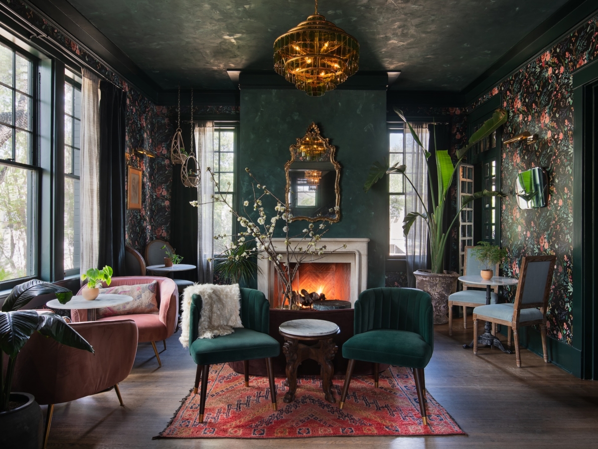 A stylish Studio 55 living space with dark floral wallpaper, green velvet chairs, a red patterned rug, and a lit fireplace. Plants and gold accents decorate the space, creating a cozy, eclectic atmosphere with natural light from tall windows.