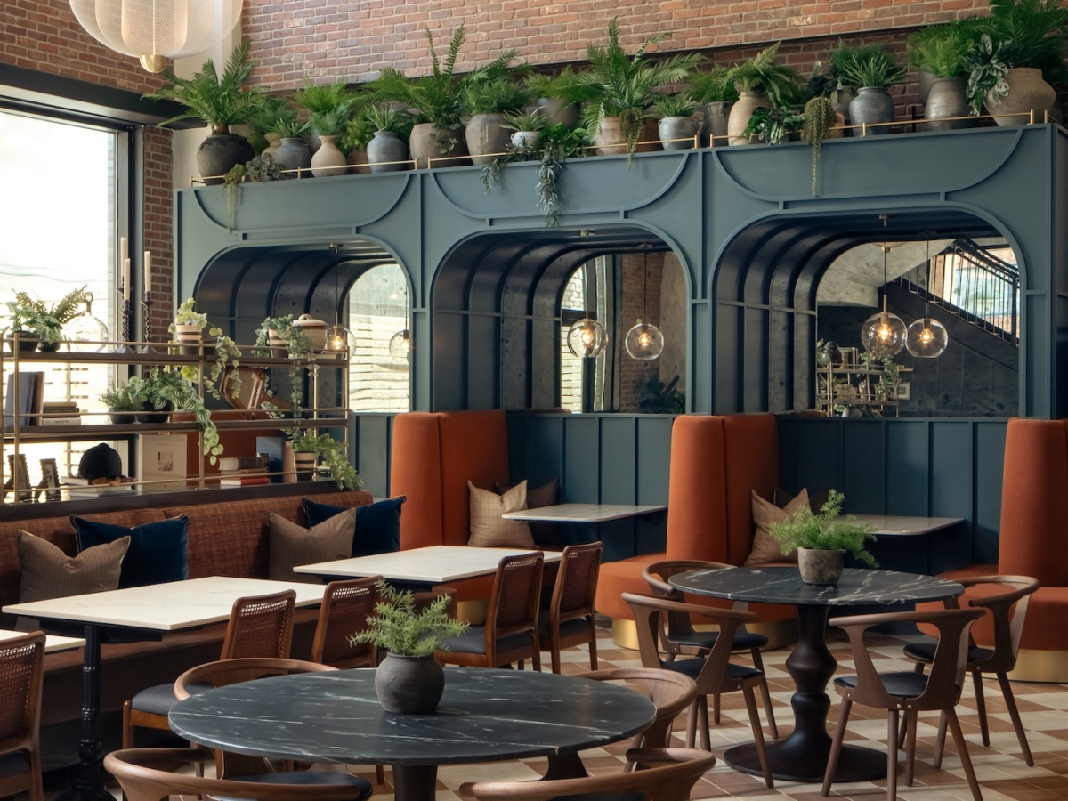 A stylish café interior by Studio 55 features round black tables, wooden chairs, cushioned seating, and a tall brick wall. Shelves with potted plants and large windows fill the space with natural light, creating a modern and inviting atmosphere.