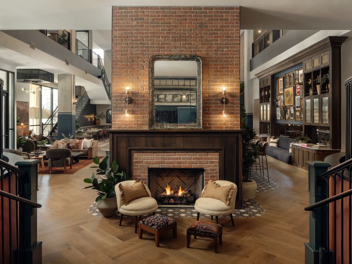 A stylish student apartment lobby by Studio 55 features a central brick fireplace, two armchairs, small ottomans, and cushions. A large mirror and sconces above the mantle add modern decor to the wood floors and open, airy atmosphere.