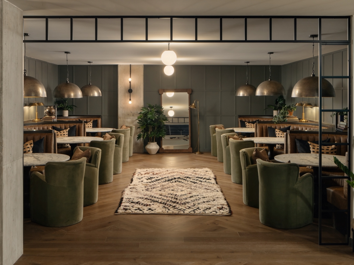 A stylish Studio 55 study mezzanine with green velvet chairs, marble tables, brown leather seating, large dome pendant lamps, indoor plants, and a patterned rug on wooden floors, creating a cozy and modern atmosphere.