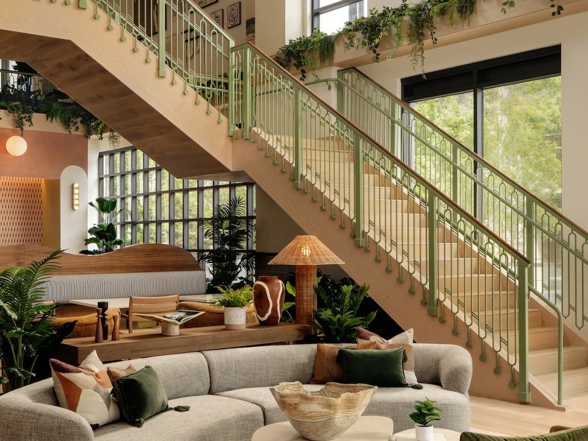 A modern apartment lobby by Studio 55 with large windows, a curved grey sofa, green accents, potted plants, light wood furniture, and a staircase with green railings. Natural light fills the room, highlighting cozy and stylish decor.