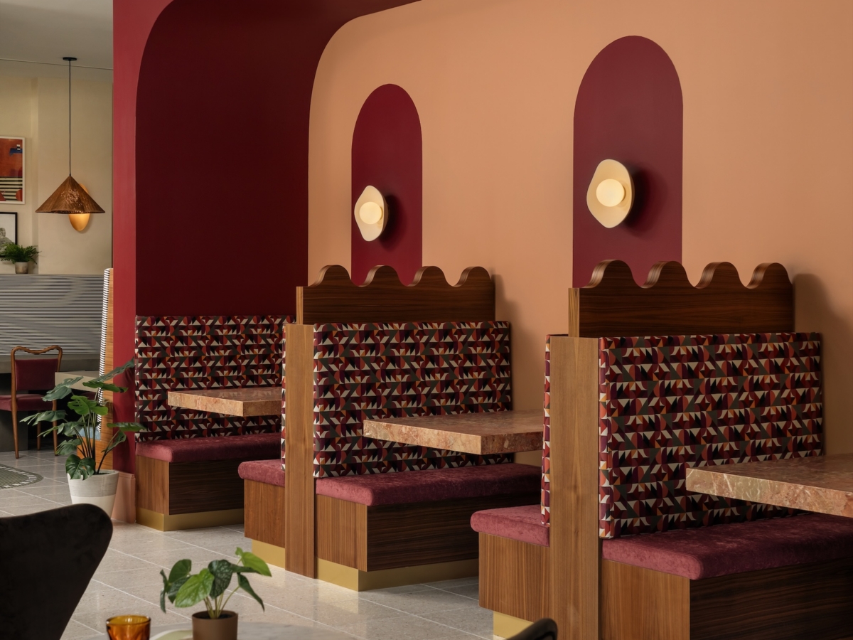 A retro-style cafe, Daydreamer, interior by Studio 55, with patterned booth seating, red and peach walls, round wall lights, and mid-century modern decor. A small table with chairs and plants is in the foreground.