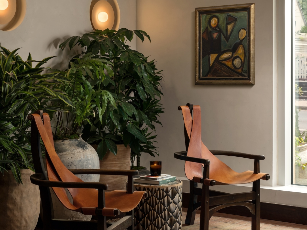Two leather chairs with wooden frames face a small round side table in an apartment common area by Studio 55. Potted plants, wall art, and round wall lights decorate the space, with a large window letting in natural light.
