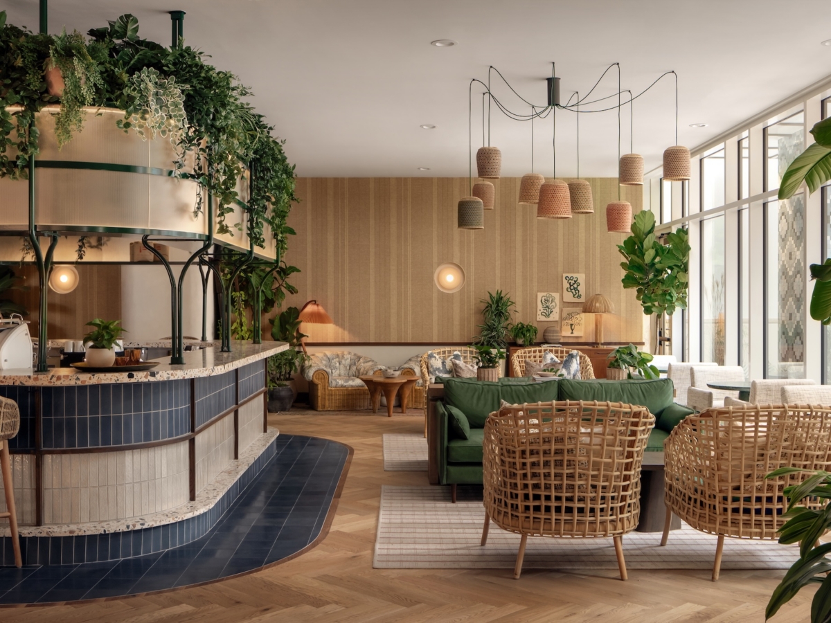 A cozy café, Daydreamer, with wicker chairs, green sofas, potted plants, woven pendant lights, and a curved counter. Large windows fill the space with natural light for a warm, inviting atmosphere rich in natural tones and greenery.
