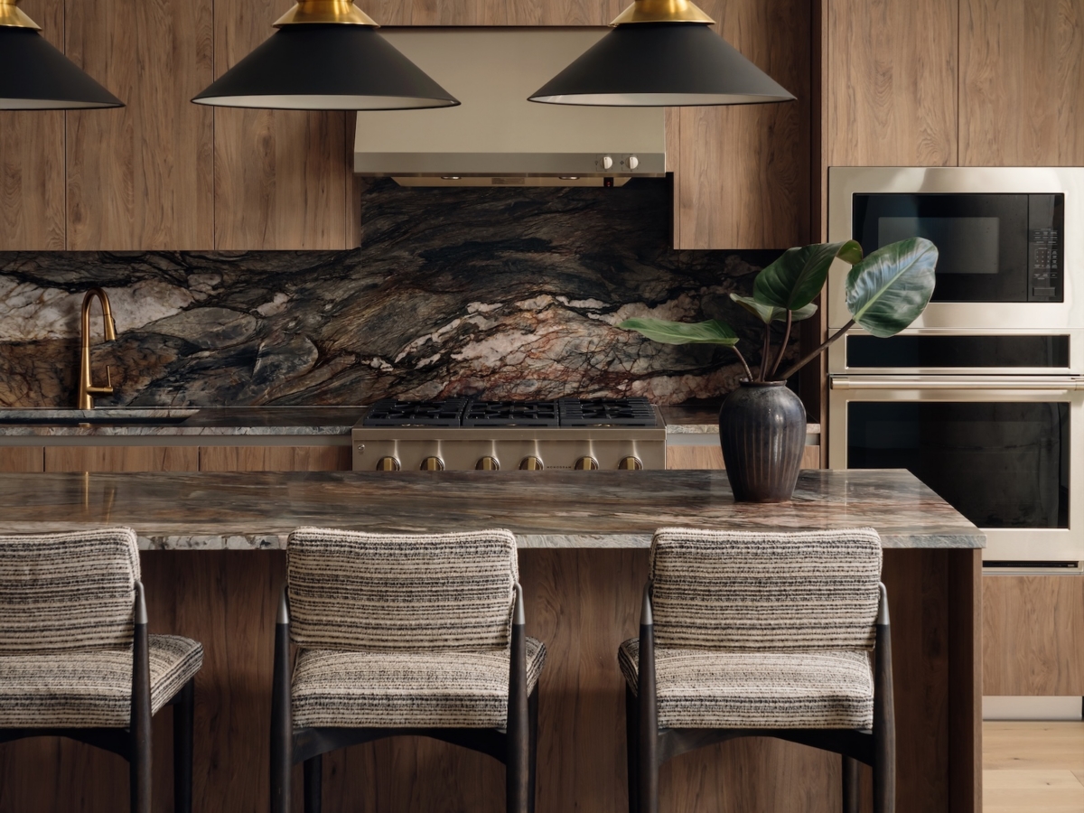 A modern Studio 55 penthouse kitchen with wood cabinetry, a marble backsplash and countertop, black and gold pendant lights, three upholstered barstools, built-in oven and microwave, and a potted plant on the island.