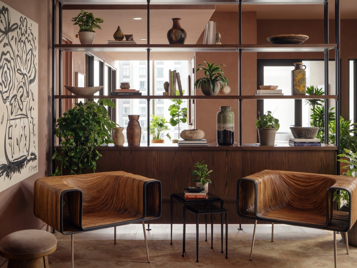 A modern shared apartment space by Studio 55 features two wooden armchairs, a black side table, green plants, pottery on open shelves, large windows, and a black-and-white abstract floral painting on a brown wall.