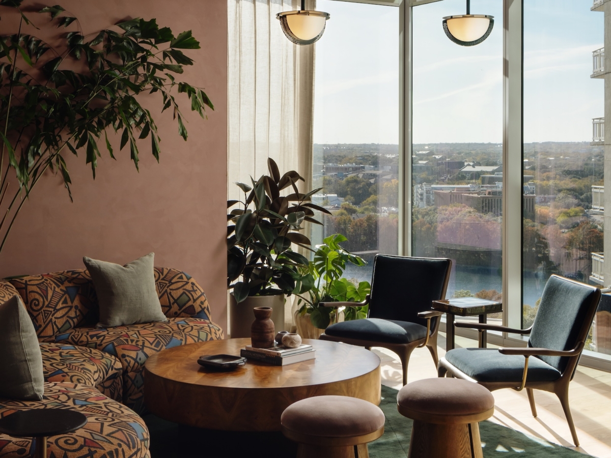 A modern Studio 55 apartment coworking space with large windows, patterned sofa, round wooden coffee table, green rug, two wooden stools, blue armchairs, tall plants, and a unique ceiling light fixture, filled with natural sunlight.
