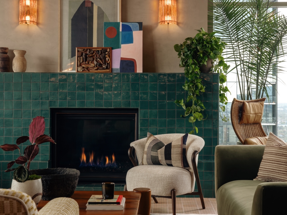 A stylish Studio 55 shared apartment space with a green tiled fireplace, abstract art, plants, cozy chairs, a green sofa, patterned rug, and a coffee table with a mug. Large window and wall sconces add light to the space.
