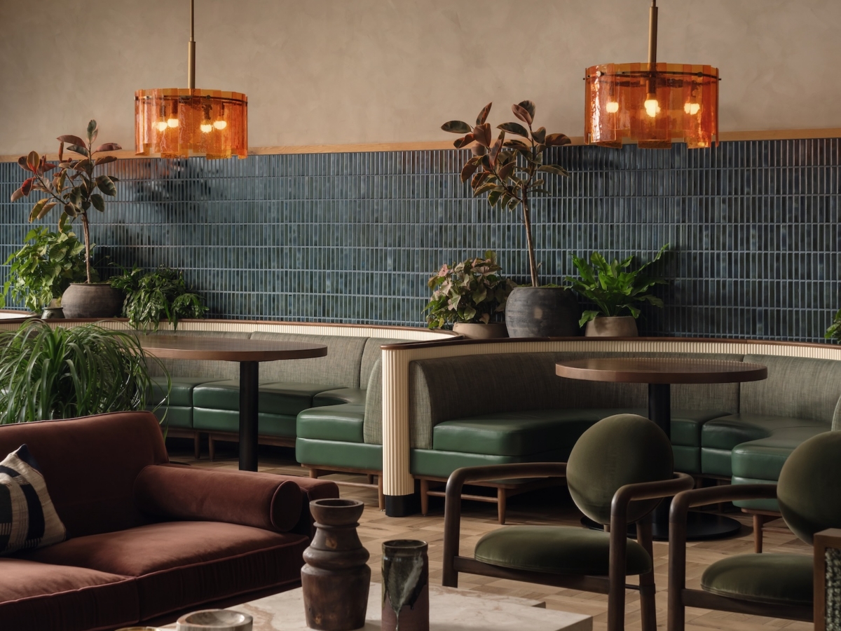 A cozy modern apartment lounge by Studio 55 with green cushioned booths, brown chairs, a marble coffee table, potted plants, and two amber pendant lights hanging from the ceiling creates a warm and inviting atmosphere.