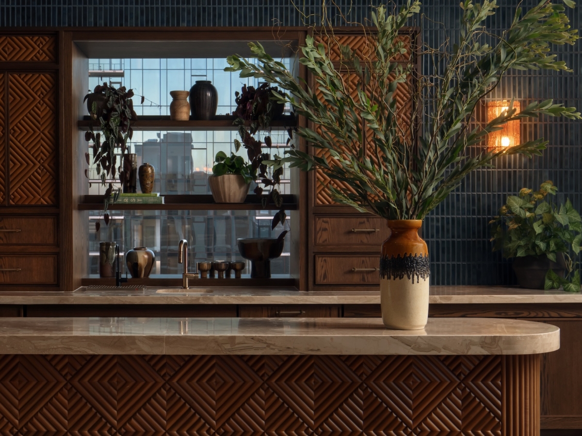 A modern shared space apartment kitchen by Studio 55 with a patterned wood island, marble countertop, and a large vase of green branches. The background features dark blue tile, wooden cabinets, shelves with plants, and decorative pottery.