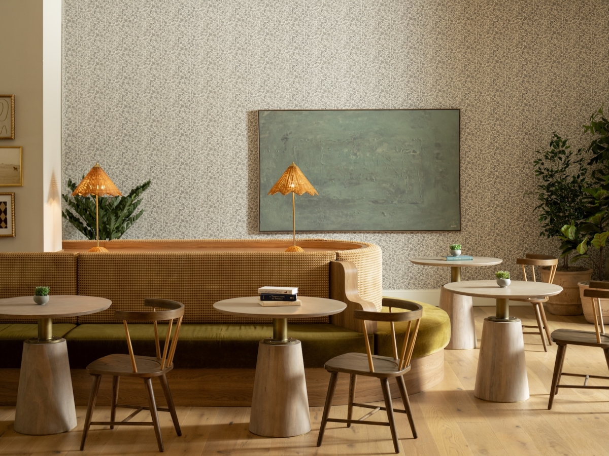 A cozy coworking space, Rambler Athens, with round tables, wooden chairs, a curved cushioned bench, small potted plants on tables, woven lamps, framed art, a large chalkboard, and green plants against a patterned wall.