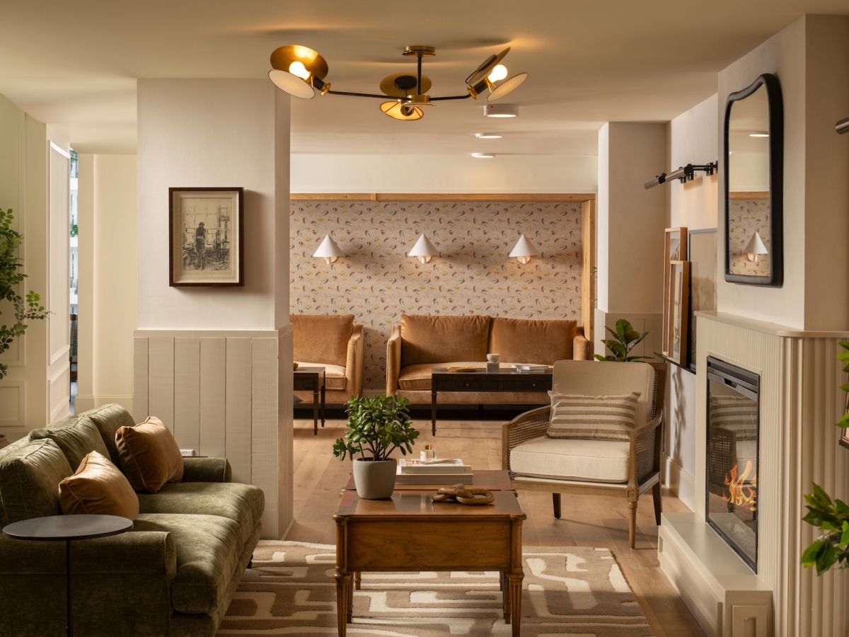 A cozy, well-lit lobby lounge by Studio 55 features a green sofa, wooden coffee table with potted plants, armchair, fireplace, and wall art. In the background are three tan chairs under wall lights and a patterned accent wall.