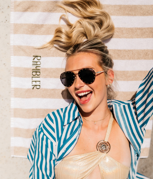 A woman with blonde hair in a high ponytail smiles while lying on a striped towel embroidered with RAMBLER by LV Collective. She wears sunglasses, a gold swimsuit, and a blue-striped shirt.