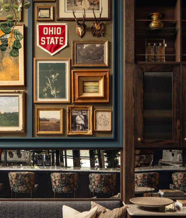 A wall gallery by Studio 55 featuring framed art, photographs, and an Ohio State pennant above a dark couch with striped pillows; a wooden cabinet and mirrored section reflect floral chairs in the background.